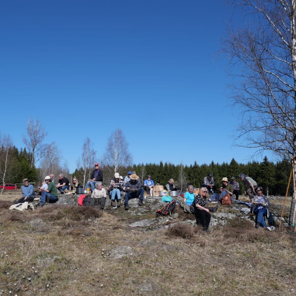 En bild med en stor gräsmatta i förgrunden, en grupp människor i mitten av bilden och direkt bakom ...